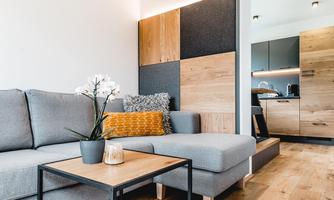 A modern living room with a gray sofa and a wooden coffee table. In the background, a kitchen with modern furniture can be seen.