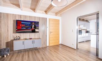 A modern living room with a flat-screen TV on the wall and a wooden shelf. The room has bright walls and plenty of daylight.