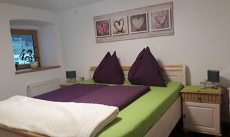 A cozy bedroom with a large bed and two purple pillows. Pictures hang on the wall and a window lets daylight in.