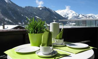 A beautiful table on a balcony with a view of the mountains. It is decorated with fresh herbs, a jug, and cups on a green cloth.