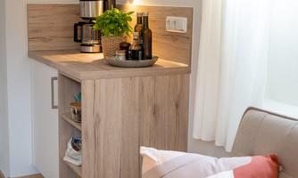 A cozy corner with a wooden cabinet, a coffee machine, and a plant. An armchair with a decorative pillow stands next to it.
