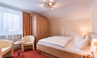 A cozy bedroom with a large bed, two armchairs, and a table. Soft colors and plenty of light create an inviting atmosphere.