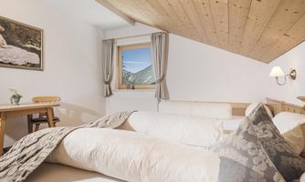 A cozy bedroom with a wooden ceiling and bright walls. There is a window with a view of the mountains and a small table.
