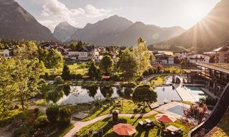 A picturesque landscape with mountains, a clear lake, and lush greenery. In the foreground, there are loungers and sun umbrellas that convey a relaxed atmosphere.