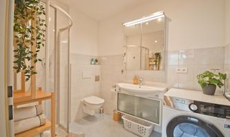 A modern bathroom with a shower, a sink, and a washing machine. Plants and towels give the room an inviting atmosphere.
