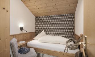 A cozy bedroom with wooden paneling and a large window. The room is simply furnished with a bed, a chair, and a small tabletop.