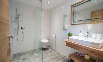 A modern bathroom with a glass shower and a stylish wooden sink. The walls are covered with white tiles and the floor is made of gray stone.