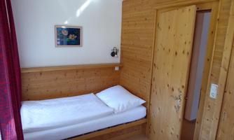 A simple room with wooden walls and a single bed. There is a window with curtains and a door that leads to another room.
