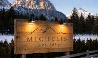 A wooden sign with the inscription "MICHELIS COTTAGE" and illumination. In the background, snow-covered mountains are visible.