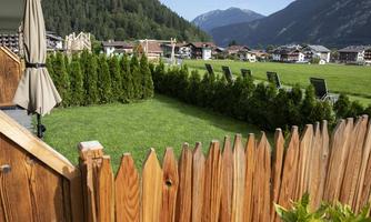 A well-kept garden with a wooden fence and green bushes. In the background, mountains and some buildings can be seen.