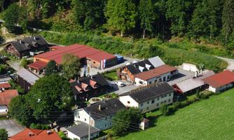 A quiet village with several buildings and red roofs, surrounded by green trees. In the foreground, a meadow can be seen.