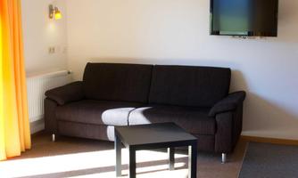 A modern living room with a gray sofa and a small table. On the wall hangs a television, and there are large windows with orange curtains.