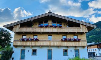 A traditional alpine house with wooden cladding and balcony flowers. In the background, gentle hills and a blue sky are visible.