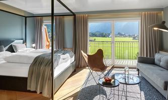 A modern hotel room with a large bed and a cozy living area. Large windows offer a view of the green landscape.
