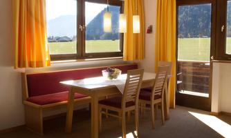 A cozy dining area with a wooden table and chairs. Large windows with yellow curtains offer a view of the landscape.