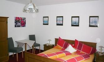 A cozy bedroom with a double bed and colorful cushions. There is a small table and chairs as well as pictures on the wall.