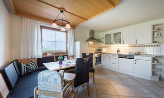 A bright and modern kitchen with a white kitchen cabinet and a dining area. The room has large windows and cozy seating options.