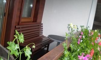 A cozy balcony seating area with a wooden bench and a small table. Colorful flowers are blooming around the balcony.