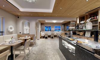 A modern restaurant with bright wooden furniture and large windows. It features an appealing breakfast bar with fresh foods.