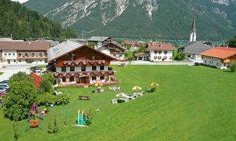 A picturesque village with traditional buildings in the foreground and majestic mountains in the background. The meadow provides a play area for children and cozy seating.