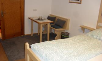 A simple room with a bed and a small table. There is a seating area and a wooden door with a warm tone.