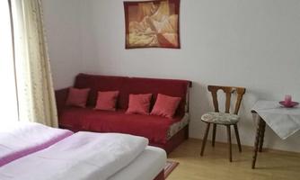 A cozy room with a double bed and a red couch. There is a small table with a chair and a piece of art on the wall.