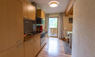 A bright kitchen with modern wooden cabinets and refrigerator. Through the window, a balcony with a view of the landscape is visible.