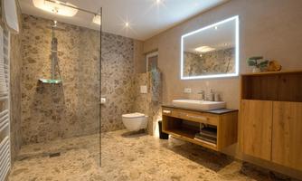 A modern bathroom with a clear glass shower and a wooden dresser. The walls are clad with textured stone, and there is a stylish, illuminated mirror.