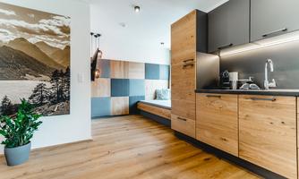 A modern kitchen with wooden cabinets and a cozy seating area. On the wall hangs a large photo of mountains and a lake.