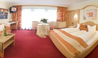 A bright, cozy hotel room with two beds and a red carpet. There is a couch, a table, and large windows with plenty of daylight.