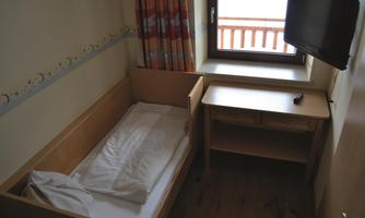 A simple room with a children's bed, a table, and a television. The window offers a view outside to a balcony.