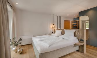 A modern bedroom with a large bed and light wooden flooring. The decor is minimalist and inviting, with soft colors and natural light.