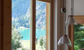 A view through a large window of a calm lake and surrounding mountains. The appearance conveys a peaceful and nature-close atmosphere.