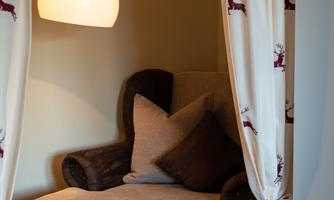 A cozy reading nook with a beige armchair and several cushions. A large, round lamp illuminates the room, surrounded by curtains with a subtle pattern.
