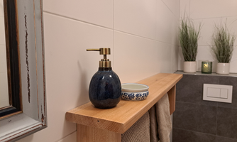 A modern bathroom shelf made of wood with a soap dispenser and a small bowl. In the background, decorative plants and a beautiful mirror are visible.