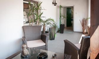 An inviting interior with rattan furniture and many plants. In the background, an open door can be seen leading to another room.