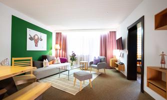 A modern living room with a sofa and a table. There are large windows, a rug, and decorative elements on the walls.