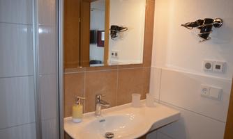 A modern bathroom with a sink, a mirror, and a shower. The walls are in neutral colors, and there are some toiletries on the shelf.