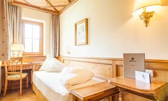 A bright room with wooden paneling and a window that lets in daylight. There is a bed, a table with a chair, and attractive decoration.