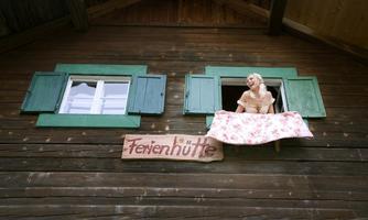 A rustic cabin with green shutters and a sign that reads "Holiday Cabin." A woman in a floral dress looks out the window.