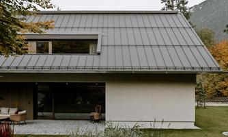 A modern house with a sloped roof and large windows. The surroundings are green and surrounded by trees.