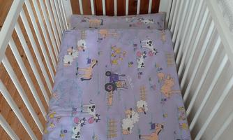 A baby bed with a white frame. The mattress is covered with a purple cover featuring cute animals and agricultural motifs.