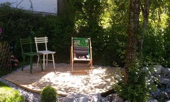 A beautiful garden with a small wooden deck, surrounded by trees and shrubs. On the deck, there is a chair and a lounge chair.