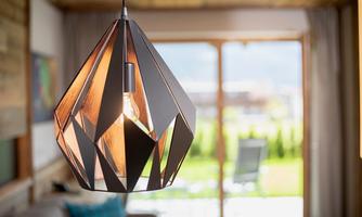 A modern lamp with an elegant, geometric design hangs over a cozy room. In the background, there is a view of a green garden and windows.