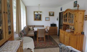 A cozy living space with a dining table and chairs. The walls are adorned with pictures, and there are wooden furniture and a carpet.
