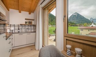 A modern kitchen corner with wooden ceilings and large windows. In the background, green meadows and mountains can be seen.