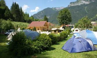 A campsite in nature with several tents and a view of green hills and mountains. In the background, a cozy wooden house is visible.