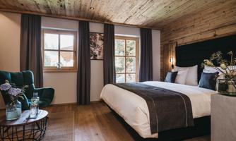 A cozy bedroom with a wooden ceiling and elegant decor. There is a large bed, a comfortable armchair, and a window with a view outside.