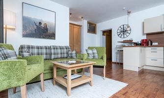 A modern living room with a green sofa and a wooden table. The open kitchen is well-equipped, and the wooden floor gives the room a cozy atmosphere.