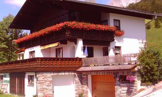 A charming house in alpine style with flower boxes and a balcony. The surroundings are green and the mountains are visible in the background.